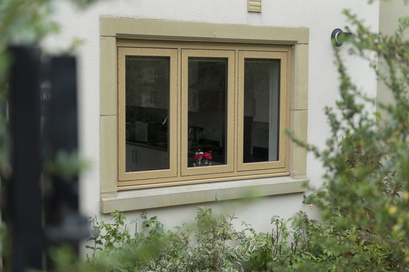 uPVC flush casement windows with light coloured woodgrain foil on a cream wall with lush green bushes in front